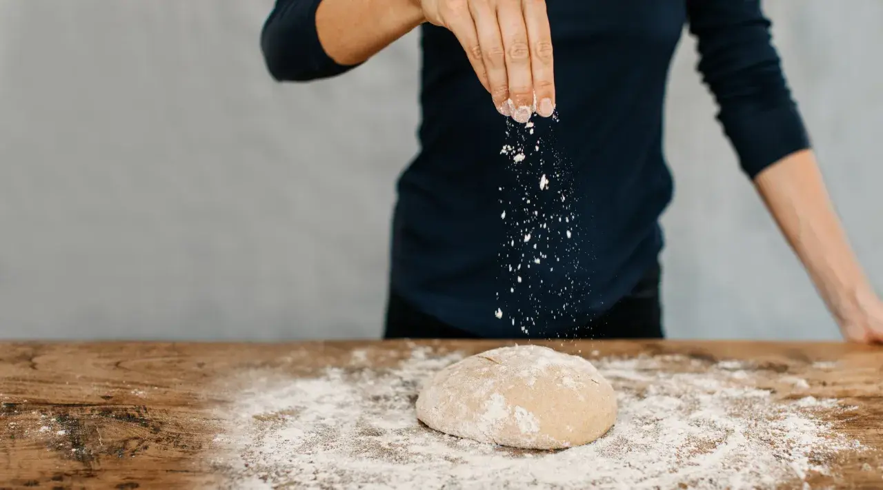 Eine Hand streut Mehl über einen Teig auf einem Holztisch. Das ist der Beginn vom backen mit glutenfreiem Mehl.