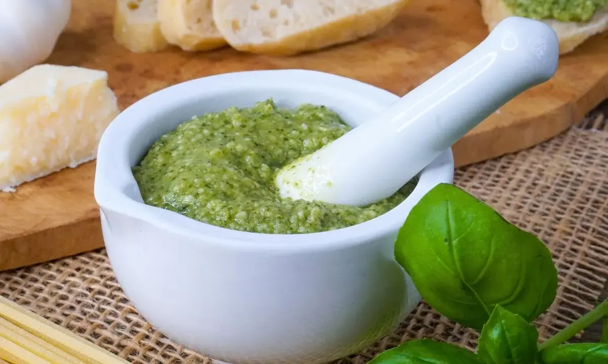 Frisches Pesto im Mörser, bereit für ein köstliches Rezept. Daneben liegen Brot, Parmesan, Knoblauch und Basilikum.