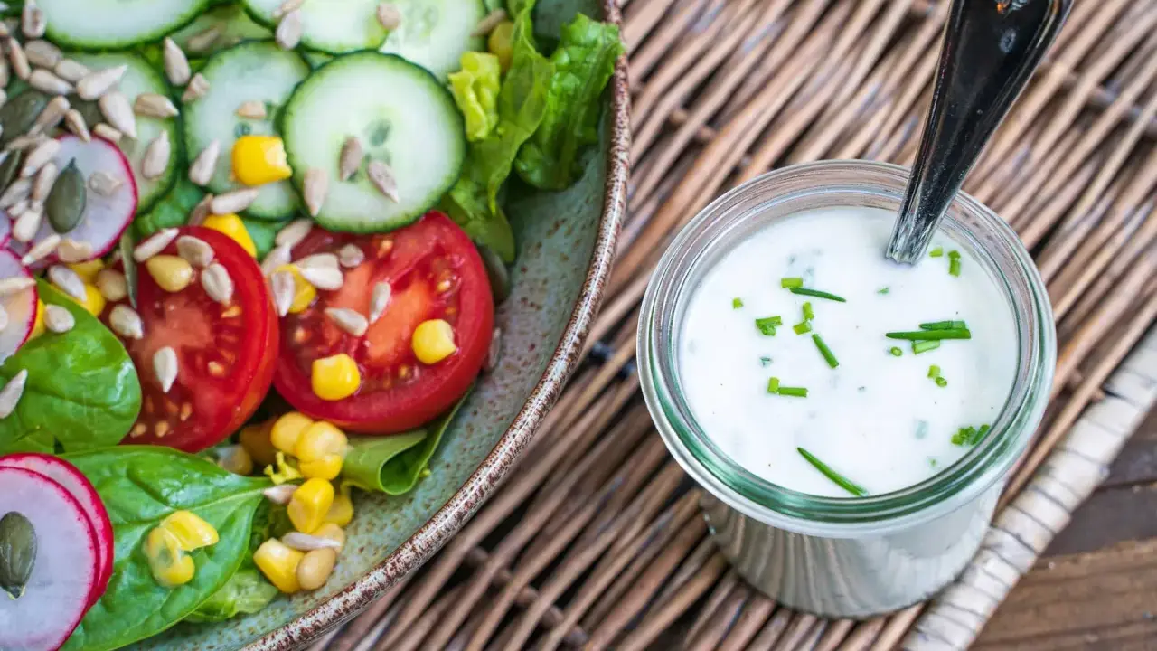 Frischer Salat mit Tomaten, Gurken und Mais, daneben ein Glas mit selbstgemachtem Joghurt-Dressing, garniert mit Schnittlauch.