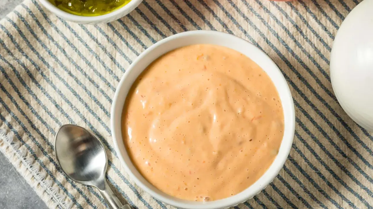 Cremige, orangefarbene Big Mac Sauce in einer Schüssel, daneben gehackte Gurken, eine Tomate und eine Zwiebel.