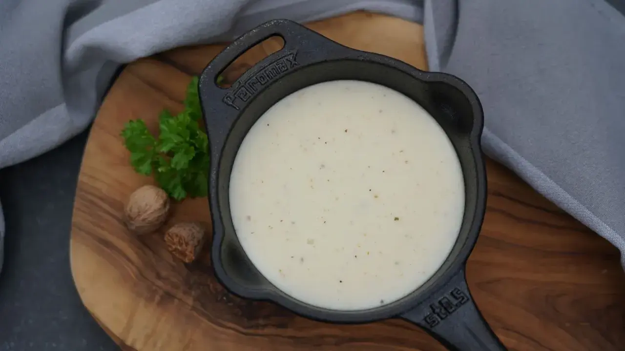 Cremige Béchamelsauce in einer kleinen Gusseisenpfanne, garniert mit Petersilie und Muskatnüssen.