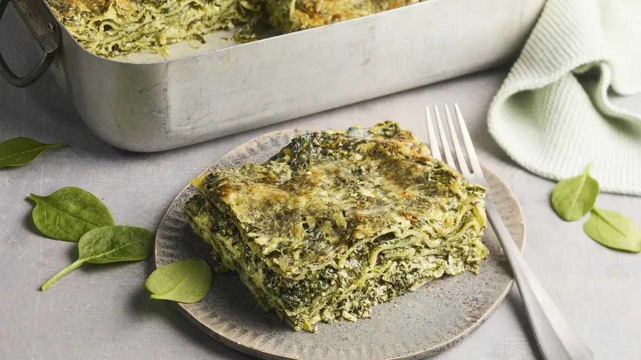 Ein Stück Spinatlasagne auf einem Teller, daneben frische Spinatblätter und eine Gabel. Das Spinatlasagne rezept ist ein Genuss!