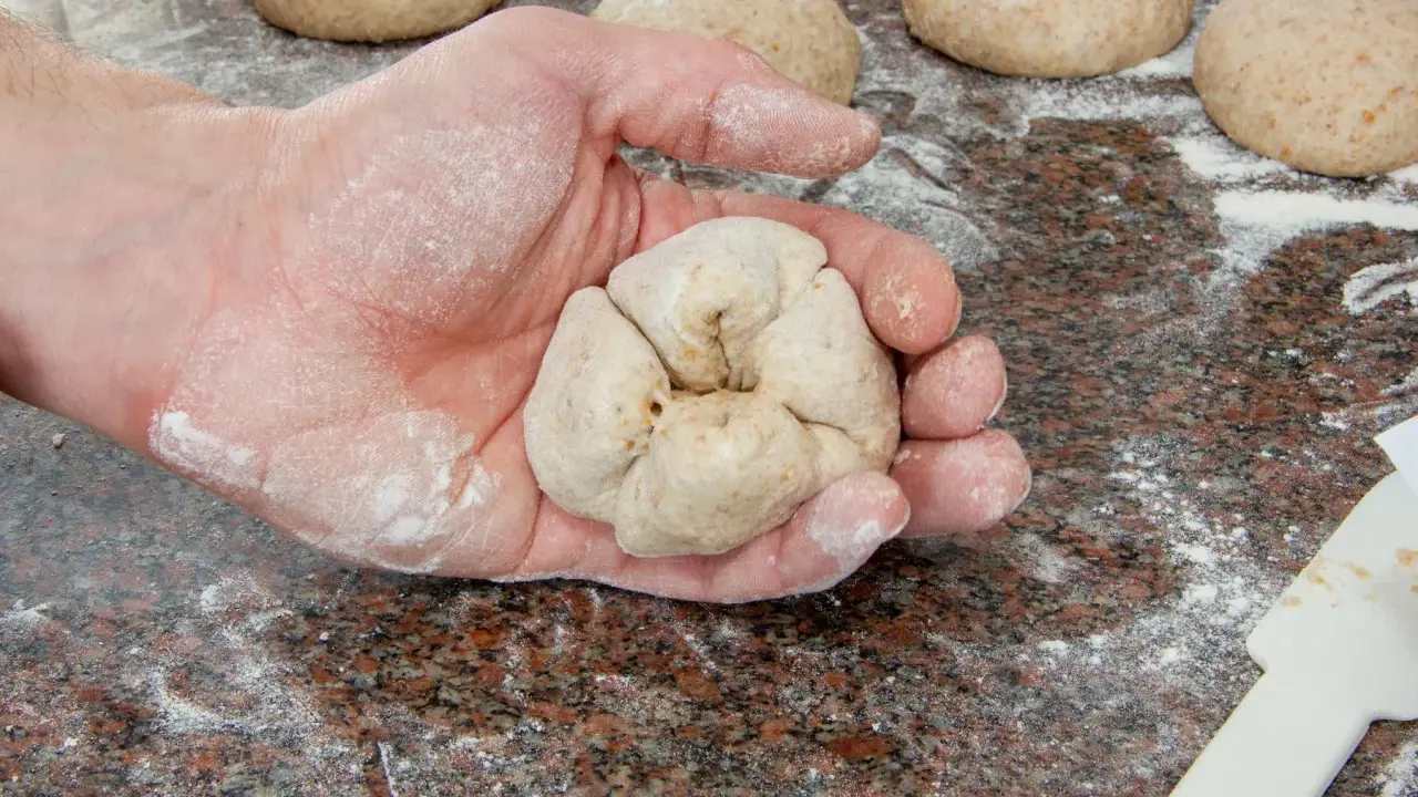 Eine Hand formt Teiglinge für das Backen mit Vollkornmehl. Mehrere Teiglinge liegen bereit auf einer bemehlten Arbeitsfläche.