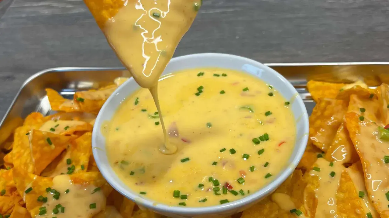 Ein Tortilla-Chip taucht in eine Schüssel mit cremiger Chili Cheese Sauce, garniert mit Schnittlauch.