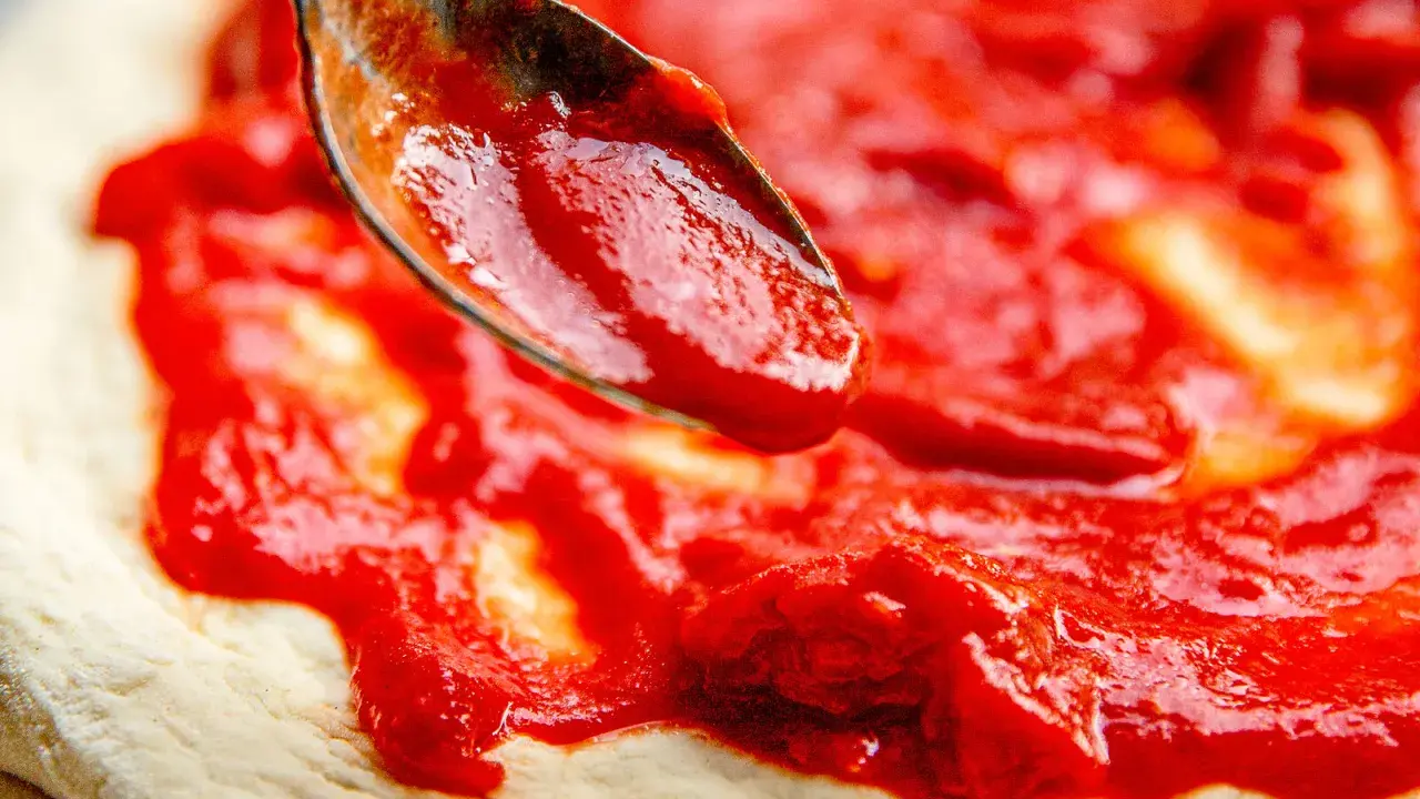 Eine Löffelspitze verteilt die leuchtend rote Pizza Sauce wie in der Pizzeria auf den Teig.