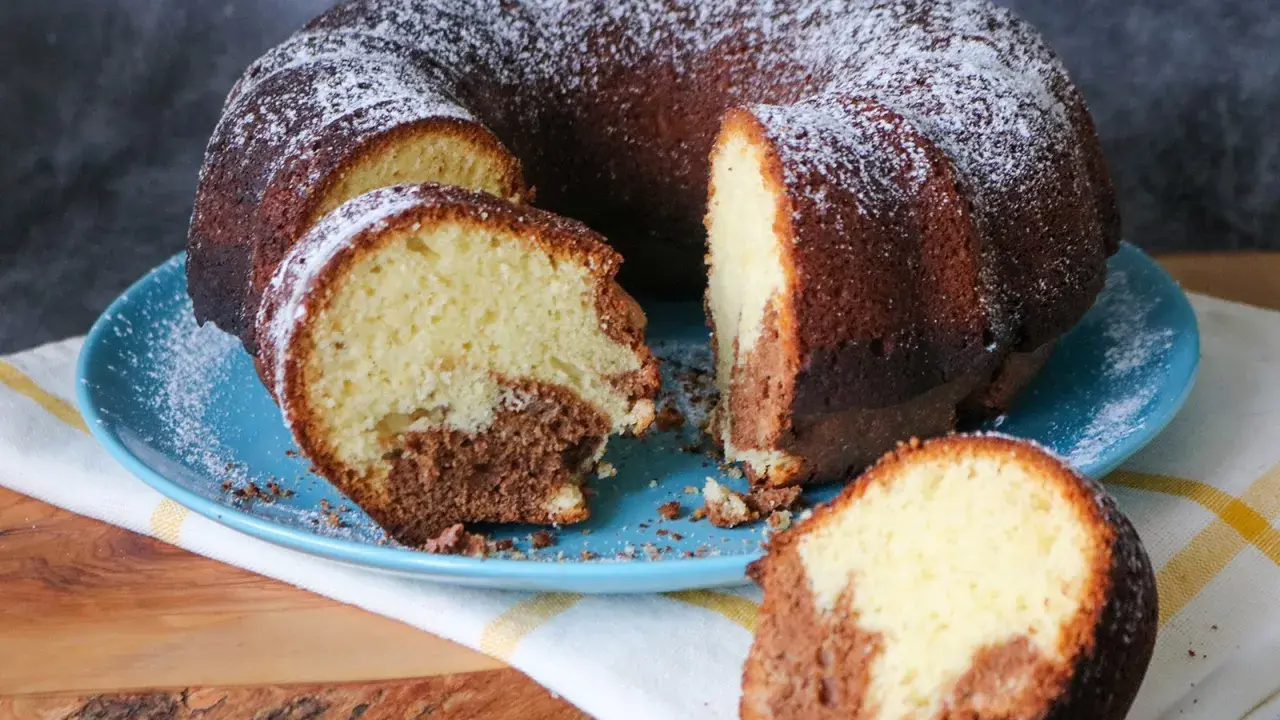 Saftiger weltbester Marmorkuchen, mit Puderzucker bestäubt, auf blauem Teller, ein Stück abgetrennt.