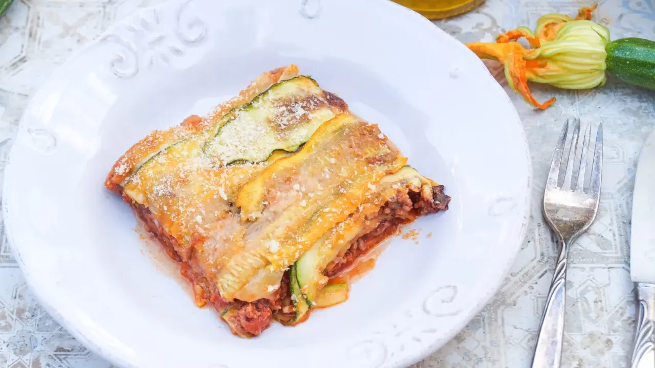 Ein Stück Zucchini-Lasagne mit Hackfleisch und Tomatensauce, bestreut mit Parmesan. Daneben liegen frische Zucchini und eine Blüte.