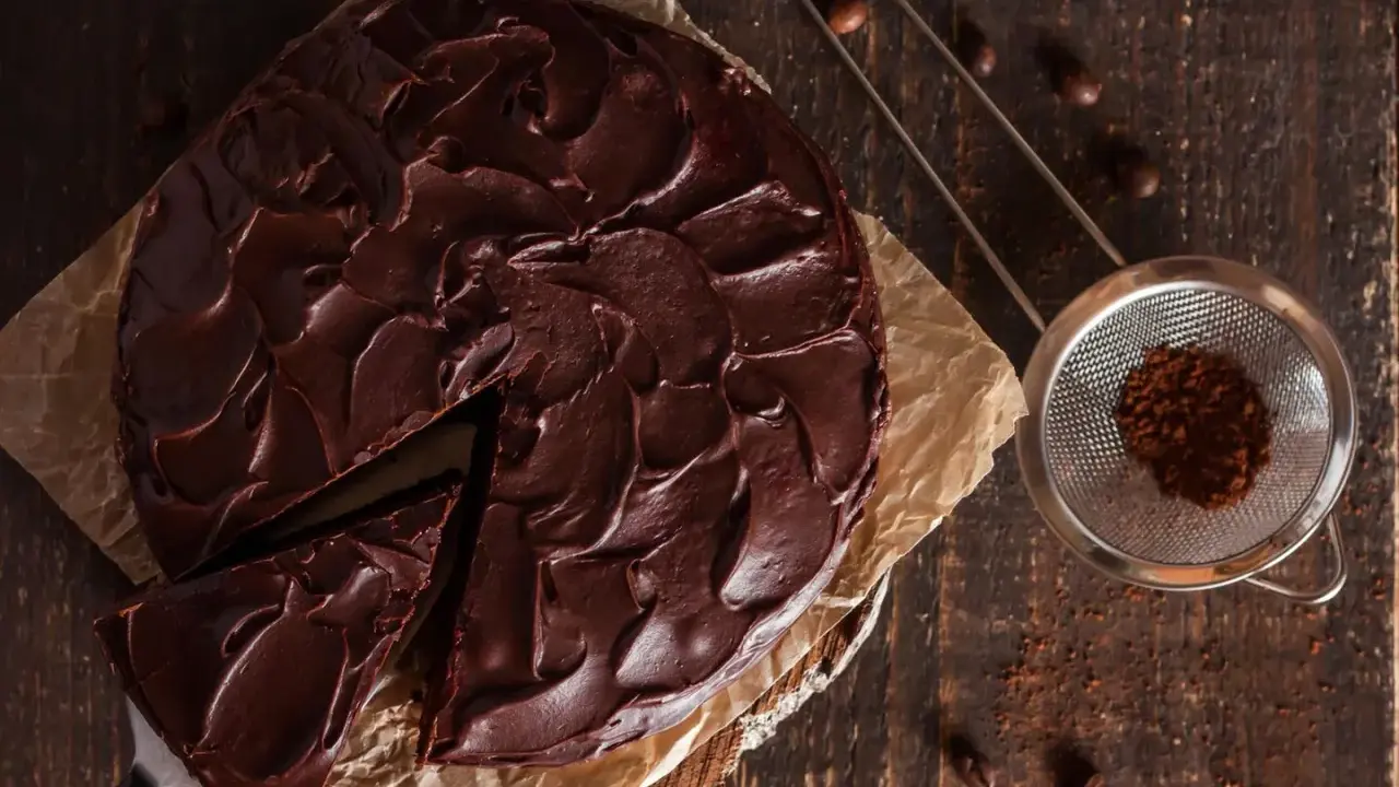 Ein saftiger Nutella Kuchen mit dunkler Schokoladenglasur, ein Stück wurde bereits entnommen. Daneben ein Sieb mit Kakaopulver.