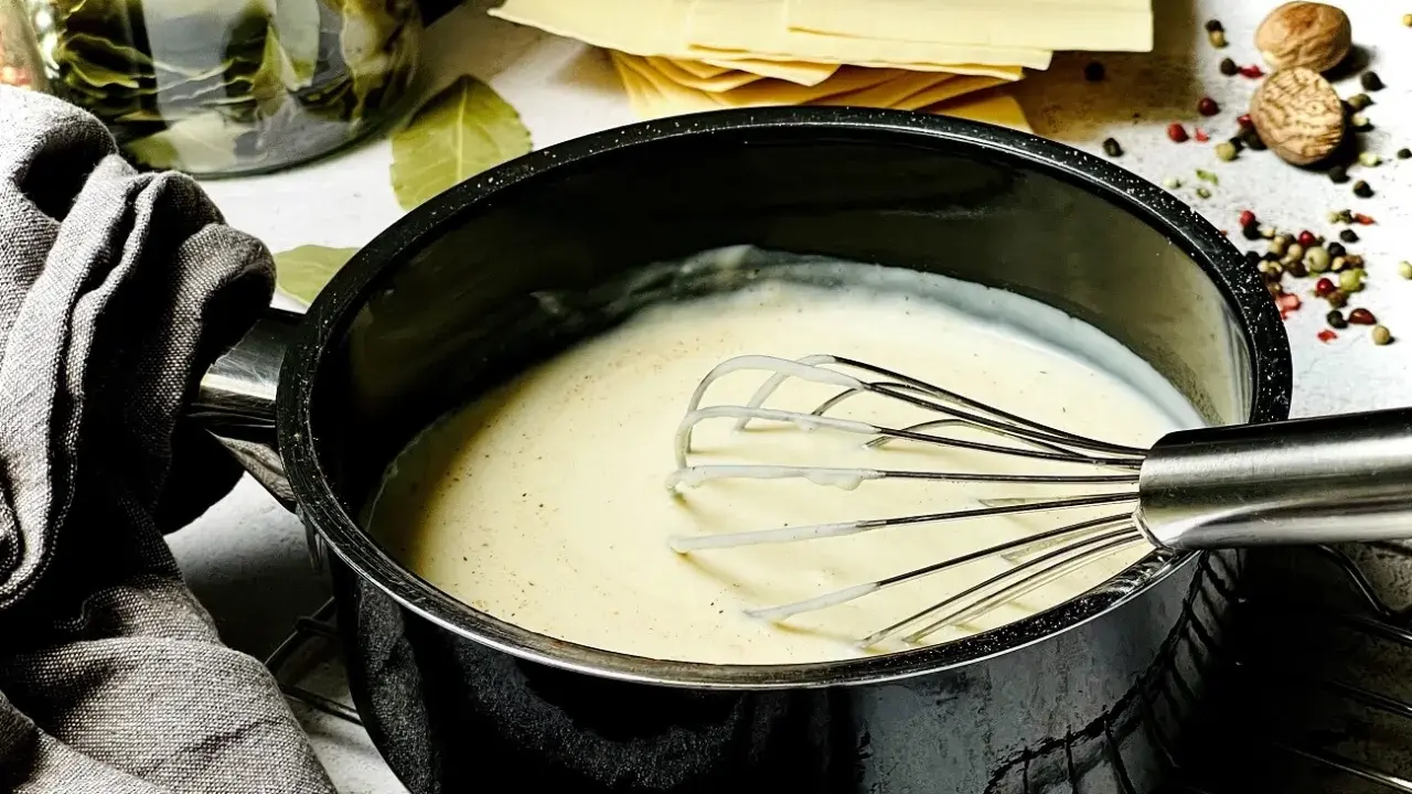Cremige Bechamelsauce in einem Topf, bereit für die Lasagne. Daneben liegen Lasagneplatten, Lorbeerblätter und Gewürze.