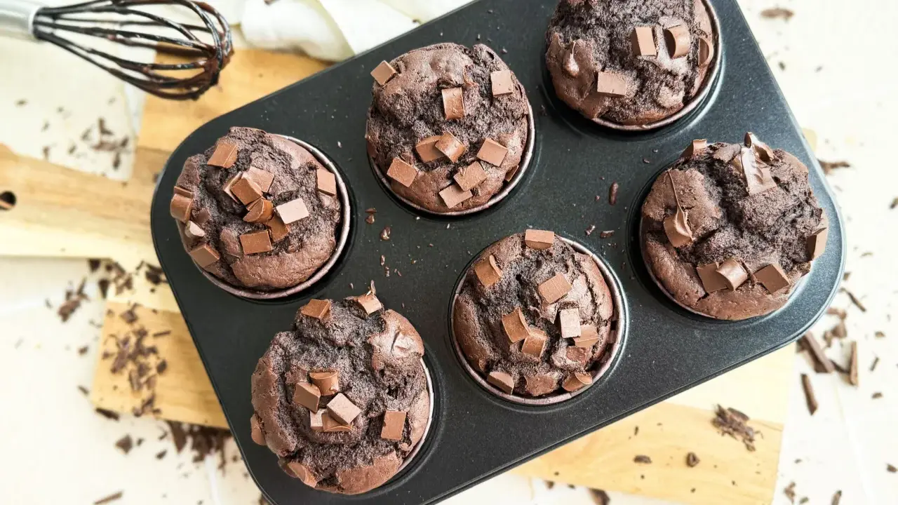 Sechs saftige Muffins mit Schokoladenstückchen in einer Muffinform. Ein Schneebesen mit Schokoladenspuren liegt daneben.