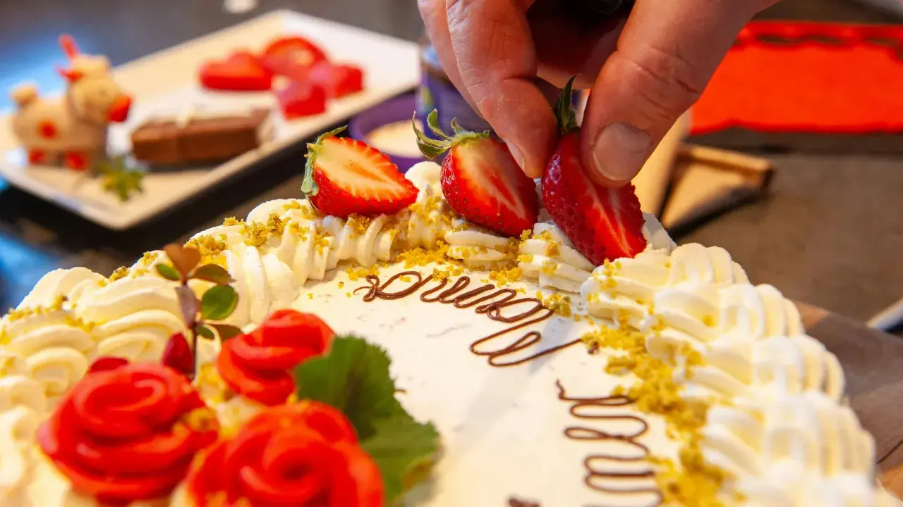 Kreative torten verzieren ideen: Eine Hand platziert Erdbeeren auf einer Sahnetorte mit Schokoladenschrift und roten Rosen.