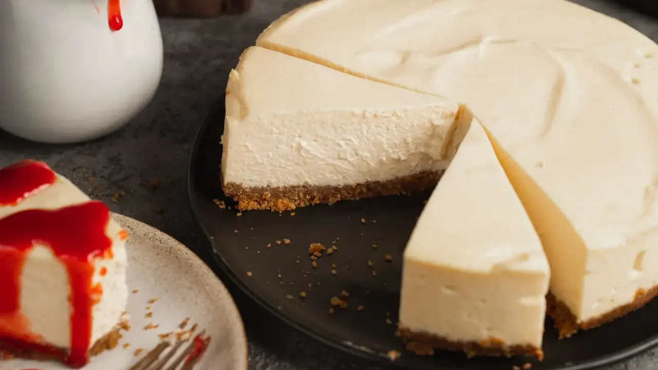 Ein cremiger american cheesecake, angeschnitten auf einem schwarzen Teller. Daneben ein Stück mit roter Soße und eine Kanne mit Soße.