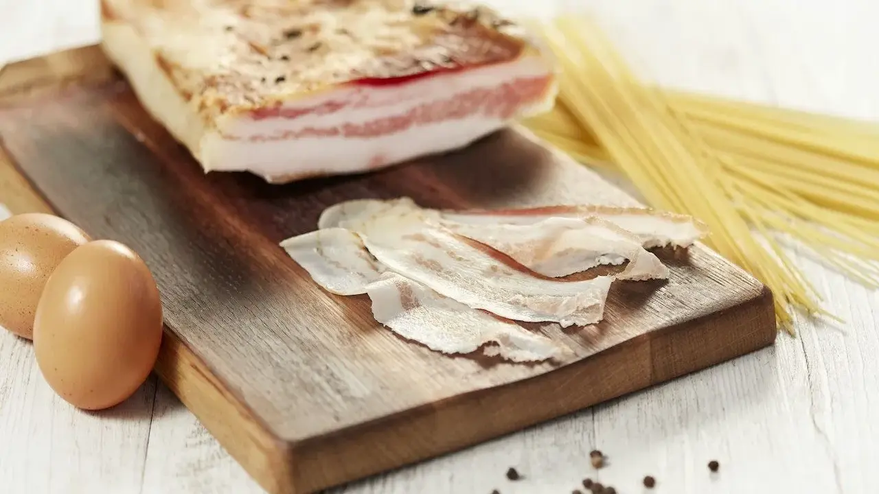 Zutaten für Guanciale Carbonara: ein Stück Guanciale, Eier, Spaghetti und Pfefferkörner auf einem Holzbrett.