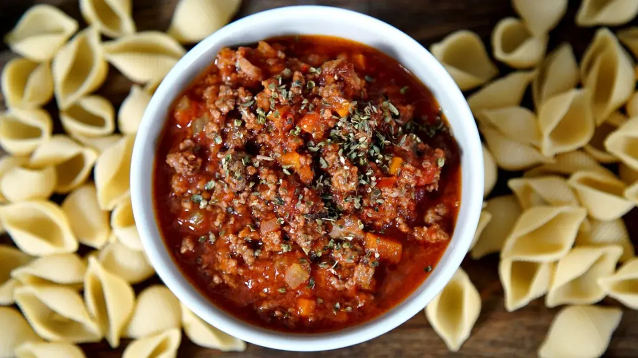 Herzhaftes Ragout alla Bolognese in einer Schüssel, umgeben von ungekochten Muschelnudeln.