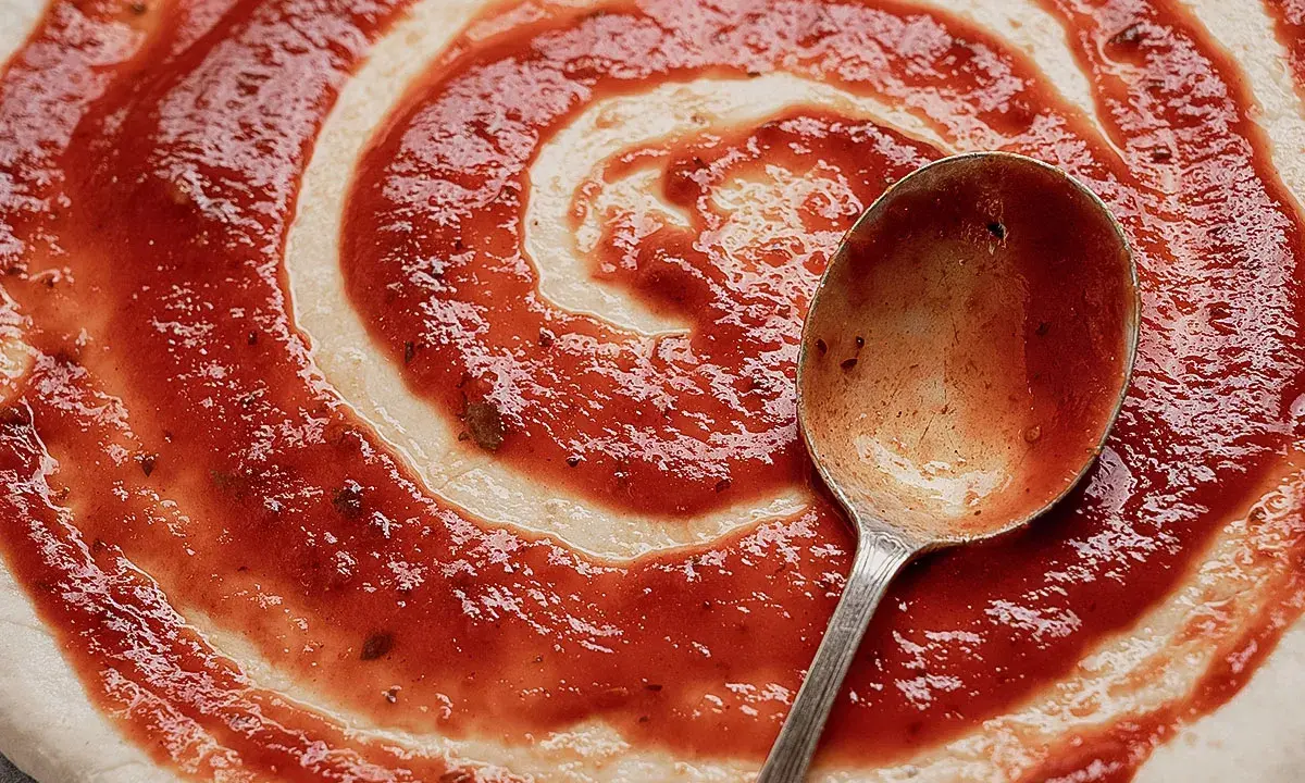 Ein Löffel verteilt das rote Pizzasaucen-Rezept auf einem Teigboden in Spiralform.