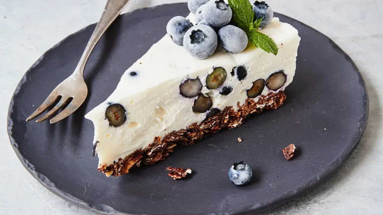 Ein Stück Heidelbeertorte ohne Backen auf schwarzer Platte, garniert mit frischen Heidelbeeren und Minze.