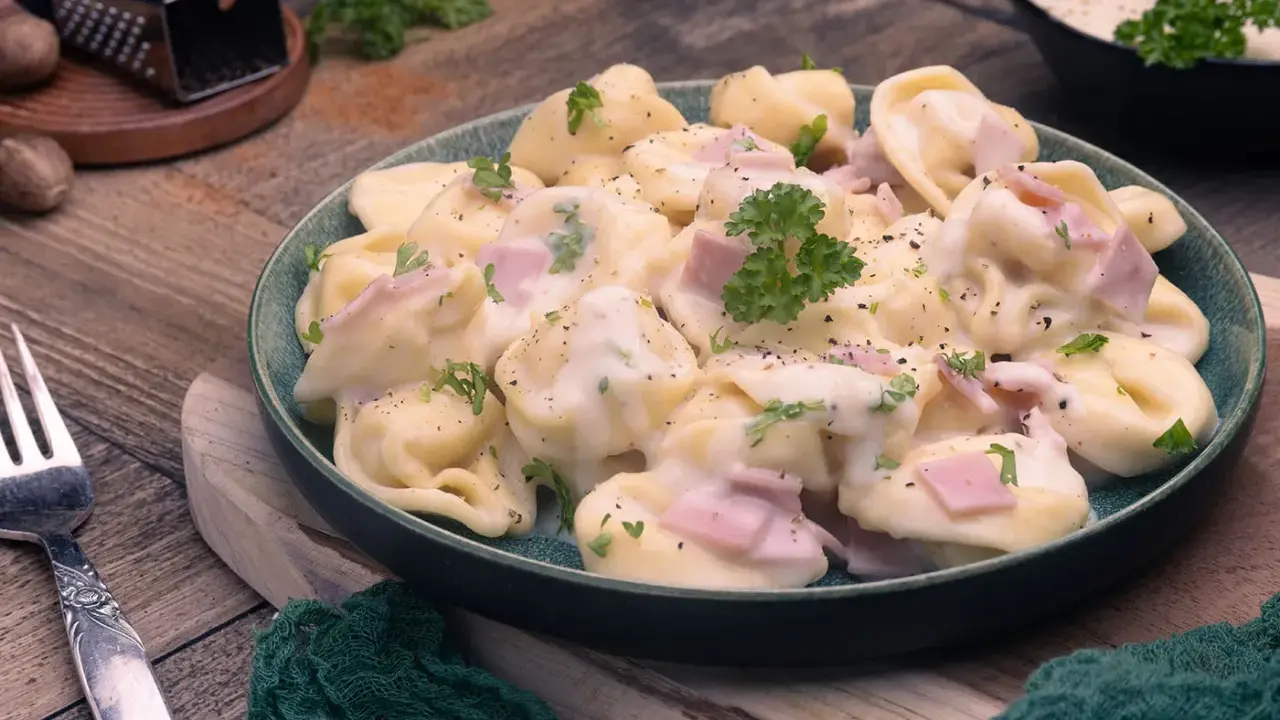 Ein Teller mit cremigen Tortellini alla Panna, garniert mit Petersilie und Schinkenwürfeln. Dazu eine Gabel und Muskatnuss.