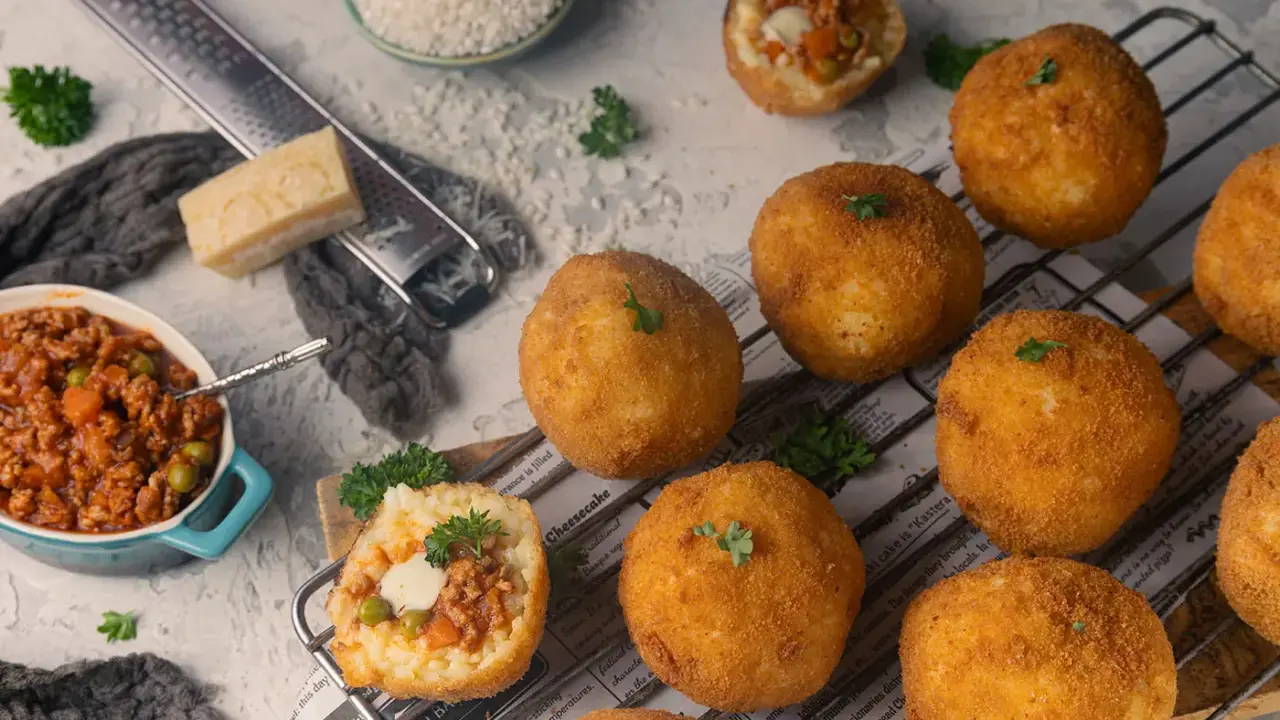Goldbraune Arancini, gefüllt mit Ragout, liegen auf einem Rost. Zutaten für das Arancini Rezept wie Reis, Parmesan und Hackfleisch sind daneben arrangiert.