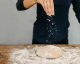 Eine Hand streut Mehl über einen Teig auf einem Holztisch. Das ist der Beginn vom backen mit glutenfreiem Mehl.