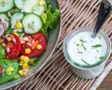 Frischer Salat mit Tomaten, Gurken und Mais, daneben ein Glas mit selbstgemachtem Joghurt-Dressing, garniert mit Schnittlauch.