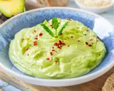 Cremige Avocado-Creme in einer blauen Schüssel, garniert mit Minze, Sesam und Chiliflocken. Daneben liegen Brotscheiben.