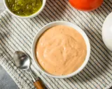 Cremige, orangefarbene Big Mac Sauce in einer Schüssel, daneben gehackte Gurken, eine Tomate und eine Zwiebel.
