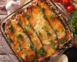 Ein Moussaka, ein Klassiker der griechischen Rezepte, in einer Auflaufform, mit geschmolzenem Käse und Petersilie garniert. Daneben liegen Knoblauch und Tomaten.