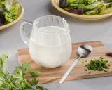 Ein Krug mit cremigem Ranch Dressing steht auf einem Holzbrett neben frischer Petersilie und Schnittlauch.