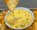 Ein Tortilla-Chip taucht in eine Schüssel mit cremiger Chili Cheese Sauce, garniert mit Schnittlauch.