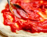 Eine Löffelspitze verteilt die leuchtend rote Pizza Sauce wie in der Pizzeria auf den Teig.