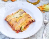 Ein Stück Zucchini-Lasagne mit Hackfleisch und Tomatensauce, bestreut mit Parmesan. Daneben liegen frische Zucchini und eine Blüte.