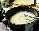 Cremige Bechamelsauce in einem Topf, bereit für die Lasagne. Daneben liegen Lasagneplatten, Lorbeerblätter und Gewürze.