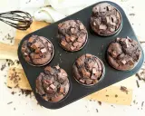 Sechs saftige Muffins mit Schokoladenstückchen in einer Muffinform. Ein Schneebesen mit Schokoladenspuren liegt daneben.