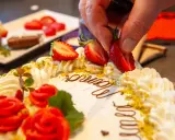 Kreative torten verzieren ideen: Eine Hand platziert Erdbeeren auf einer Sahnetorte mit Schokoladenschrift und roten Rosen.