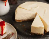 Ein cremiger american cheesecake, angeschnitten auf einem schwarzen Teller. Daneben ein Stück mit roter Soße und eine Kanne mit Soße.
