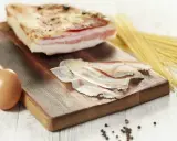 Zutaten für Guanciale Carbonara: ein Stück Guanciale, Eier, Spaghetti und Pfefferkörner auf einem Holzbrett.