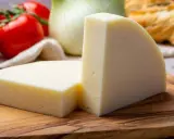 Zwei Keile hellgelben Käses, eine tolle provolone Alternative, liegen auf einem Holzbrett. Tomaten und Fenchel im Hintergrund.