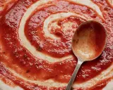 Ein Löffel verteilt das rote Pizzasaucen-Rezept auf einem Teigboden in Spiralform.