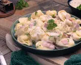 Ein Teller mit cremigen Tortellini alla Panna, garniert mit Petersilie und Schinkenwürfeln. Dazu eine Gabel und Muskatnuss.