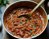 Eine Pfanne mit klassischer Bolognese, garniert mit Petersilie. Daneben liegen Knoblauch und Kräuter.