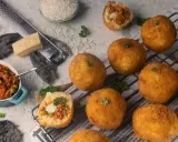 Goldbraune Arancini, gefüllt mit Ragout, liegen auf einem Rost. Zutaten für das Arancini Rezept wie Reis, Parmesan und Hackfleisch sind daneben arrangiert.