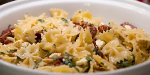Farfalle-Nudelsalat mit getrockneten Tomaten, Feta und Pinienkernen, garniert mit frischen Kräutern. Ein köstliches Gericht.