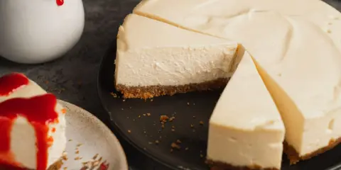 Ein cremiger american cheesecake, angeschnitten auf einem schwarzen Teller. Daneben ein Stück mit roter Soße und eine Kanne mit Soße.