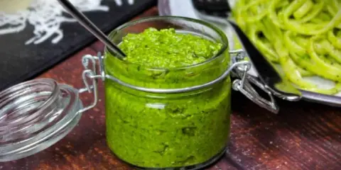 Frisches Rucola Pesto in einem Glas mit Löffel, daneben eine Portion Pasta.