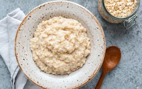 Cremiger Haferbrei im Teller, daneben ein Glas Haferflocken und ein Holzlöffel. Perfekt für ein einfaches Porridge Rezept.
