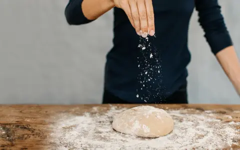 Eine Hand streut Mehl über einen Teig auf einem Holztisch. Das ist der Beginn vom backen mit glutenfreiem Mehl.