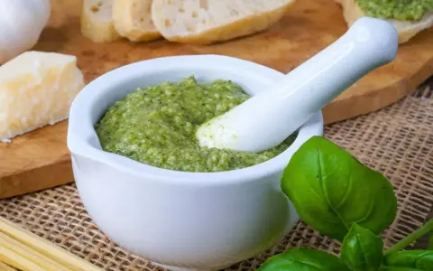 Frisches Pesto im Mörser, bereit für ein köstliches Rezept. Daneben liegen Brot, Parmesan, Knoblauch und Basilikum.