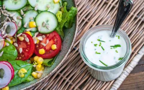 Frischer Salat mit Tomaten, Gurken und Mais, daneben ein Glas mit selbstgemachtem Joghurt-Dressing, garniert mit Schnittlauch.