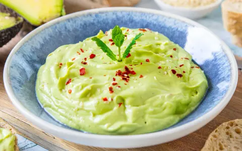 Cremige Avocado-Creme in einer blauen Schüssel, garniert mit Minze, Sesam und Chiliflocken. Daneben liegen Brotscheiben.