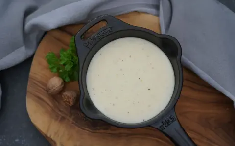 Cremige Béchamelsauce in einer kleinen Gusseisenpfanne, garniert mit Petersilie und Muskatnüssen.